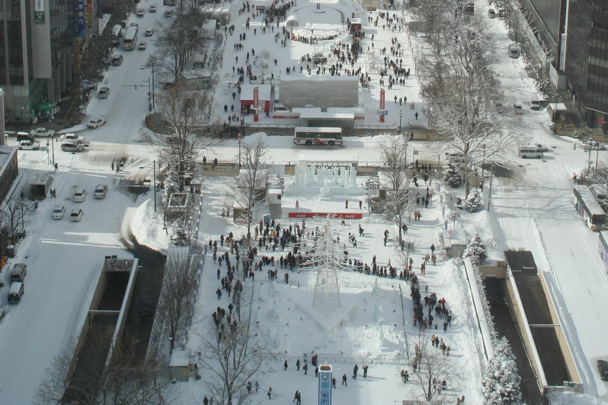 An aerial view down Odori Park during the Sapporo Snow Festival, snow sculptures lined along the park with crowds moving between them and the city and mountains beyond