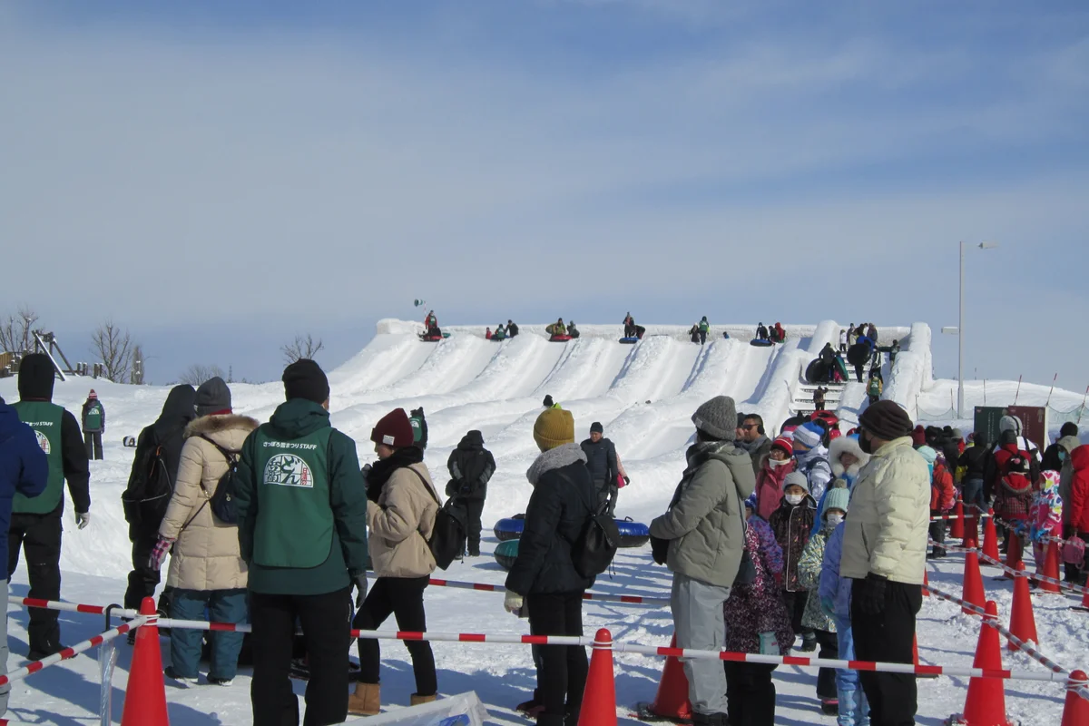 Visitors lined up at the base of a large snow slide at the Tsudome venue of the Sapporo Snow Festival, families waiting with inner tubes