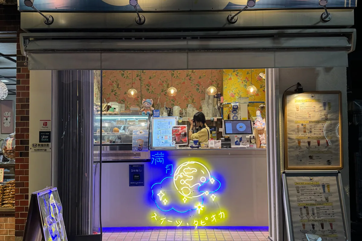 A small tapioca drink shop glowing with neon signage after dark in Shimokitazawa, a staff member working behind the counter