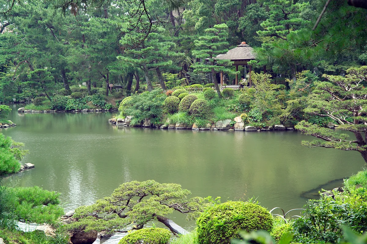 The pond and pine trees of Shukkei-en garden in Hiroshima, with a thatched-roof pavilion visible across the water