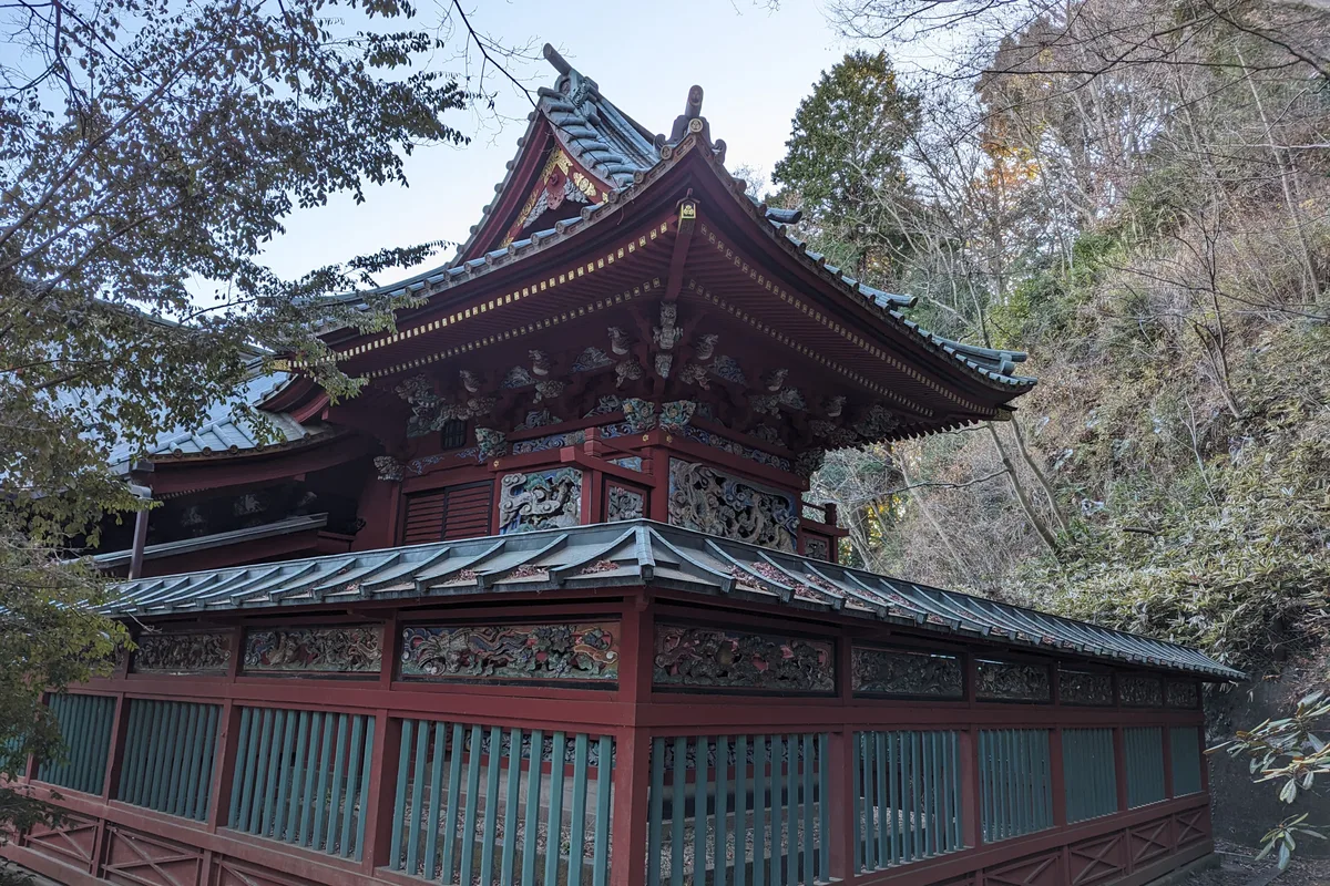 An ornate vermillion hall with carved dragon reliefs and a blue tiled roof, part of the Yakuoin temple complex on Mount Takao, surrounded by bare autumn trees