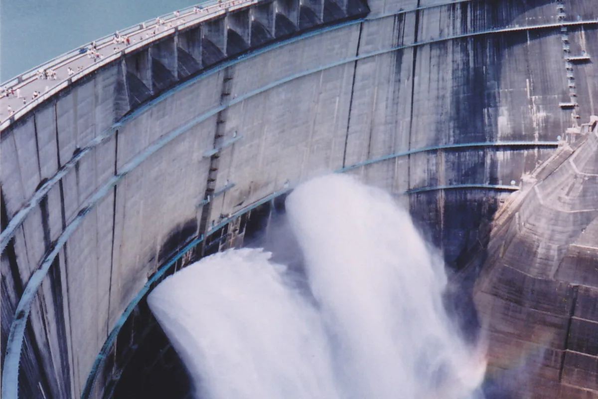 Kurobe Dam releasing a huge plume of spray water with a faint rainbow visible in the mist, visitors walking along the dam crest above