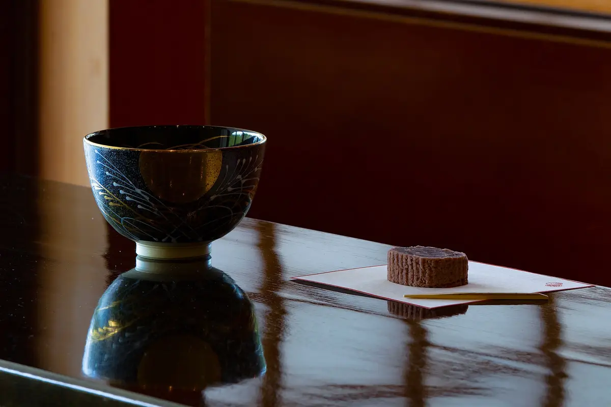 A dark tea bowl painted with a moon and grasses stands on a polished table, beside a small sweet resting on folded paper with a wooden pick