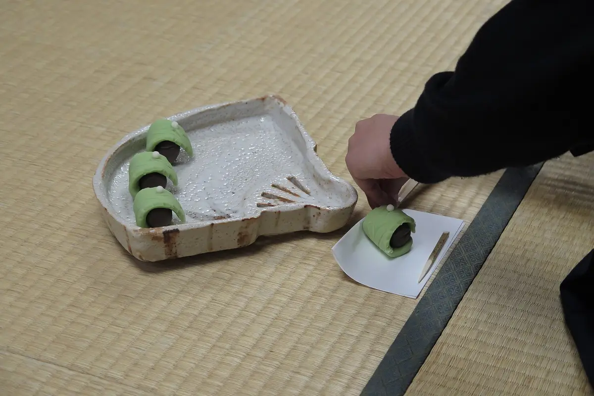A hand lifts a wooden pick away from a green sweet resting on a small folded paper on the tatami, beside a ceramic tray holding three more
