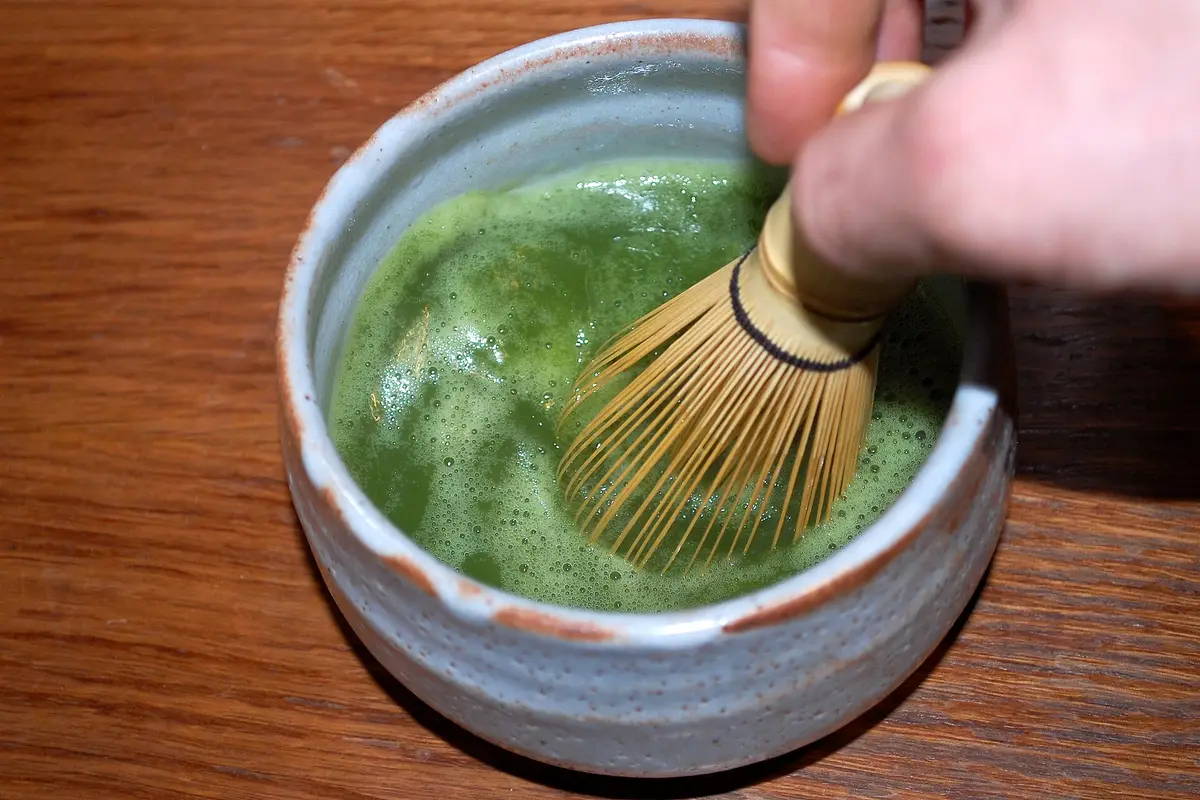 Seen from above: a hand works a bamboo whisk through bright green matcha in a pale ceramic bowl