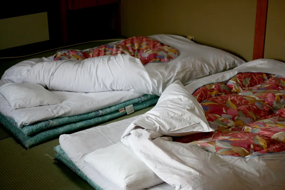 Futon bedding laid out on the tatami floor of a Japanese inn room, with white pillows beside patterned quilts