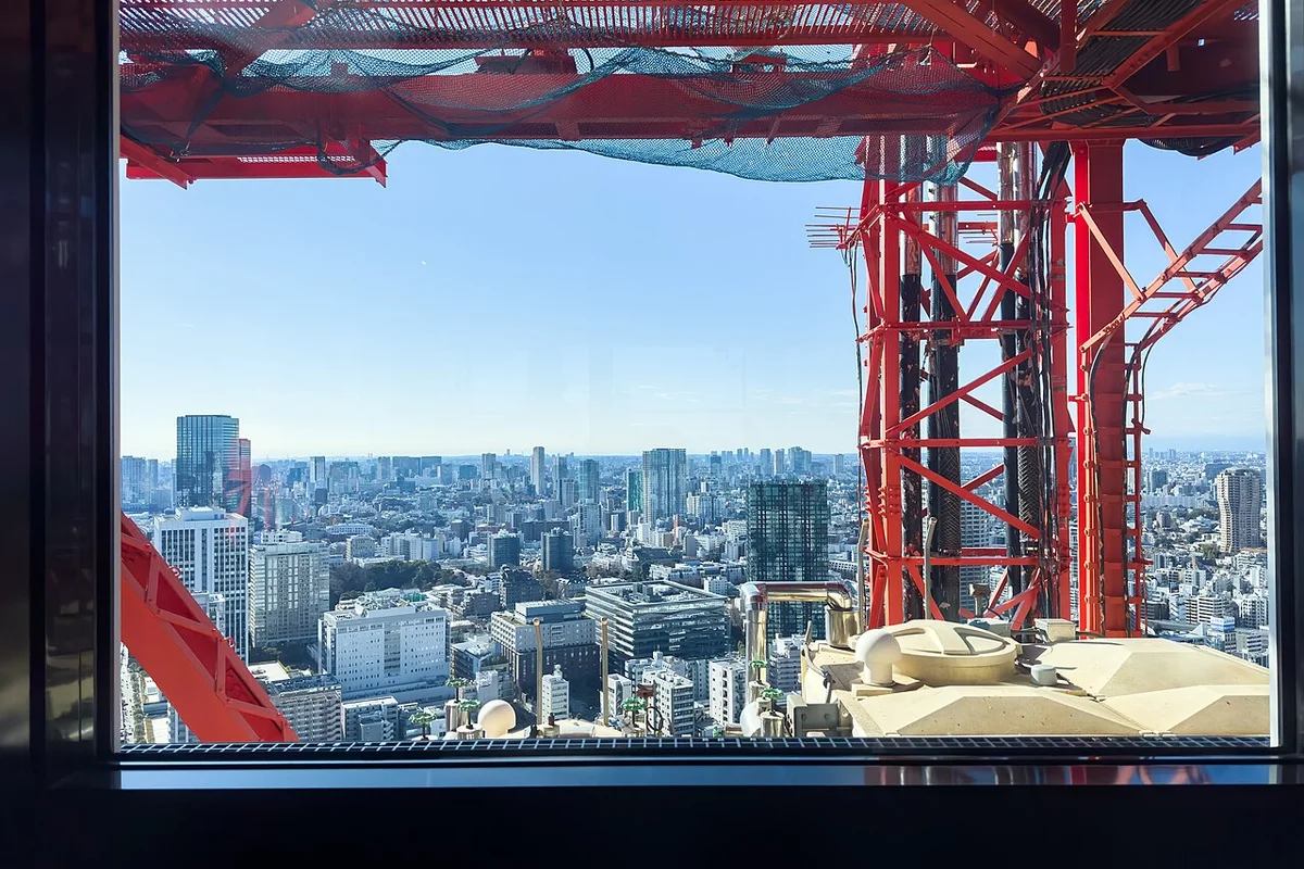 Looking out through a window near the top of Tokyo Tower, past the tower's own red steel framework and safety netting, at the dense Tokyo skyline far below