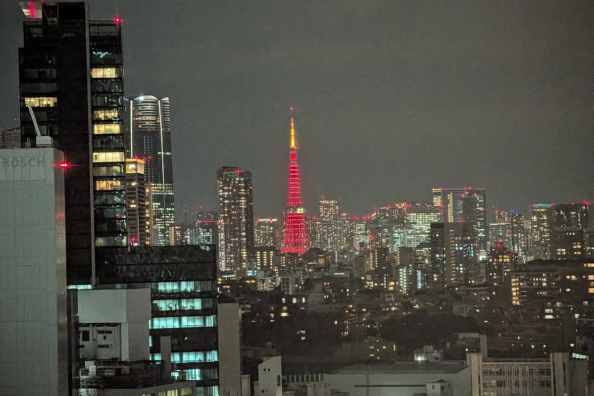 Tokyo Tower lit in red, seen from a distance and dwarfed by the dark silhouettes of surrounding high-rise towers at night