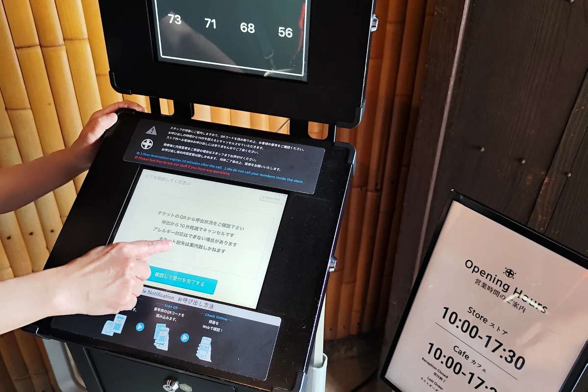 A digital queue-number display at a Uji tea house showing dozens of waiting groups still ahead, with a printed sign listing the shop's opening hours alongside it