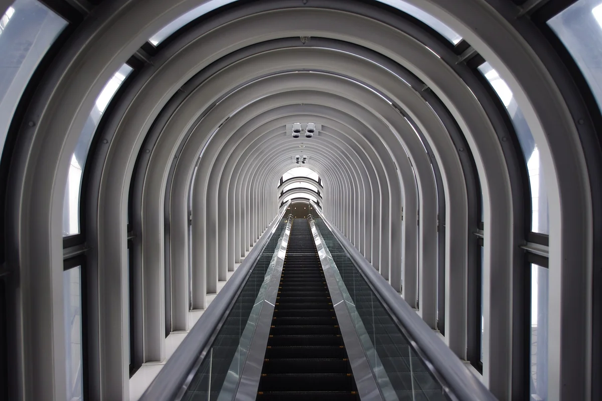 A long glass-enclosed escalator tube climbing through a series of white arched ribs toward a bright opening at the top