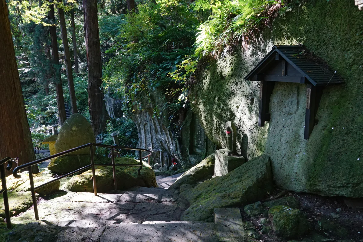 Stone steps and a handrail beside a moss-covered rock face carved with small Jizo statues along the Yamadera climb