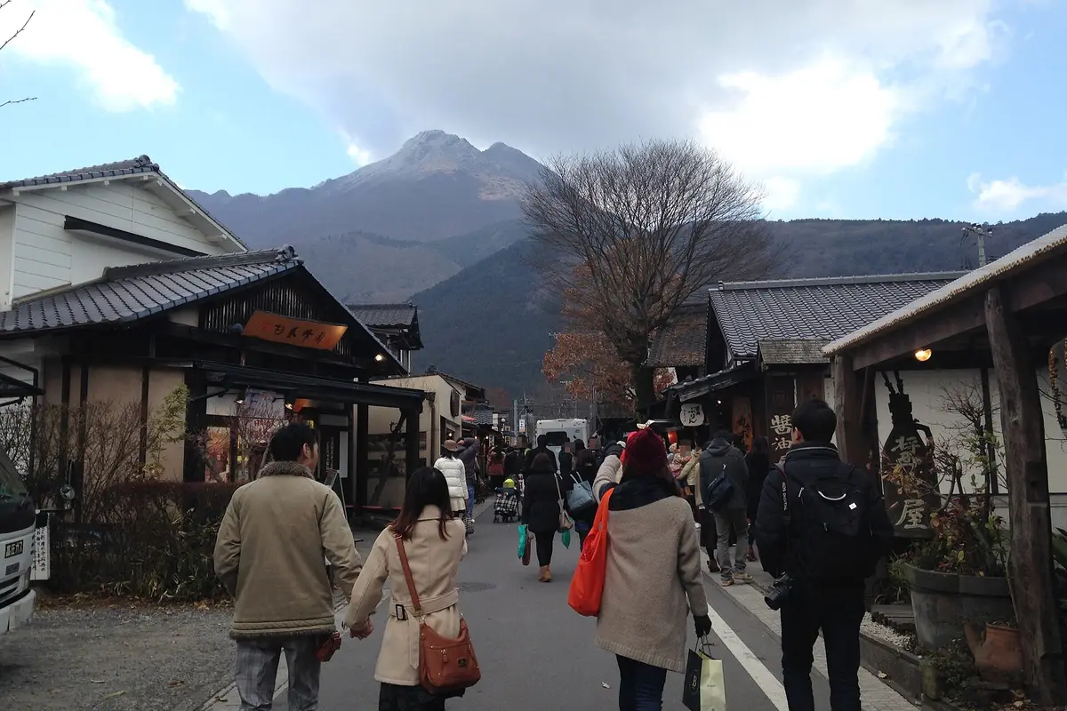 Looking up the Yunotsubo lane in Yufuin, people walking between wooden shopfronts with the twin peaks of Mount Yufu closing off the far end