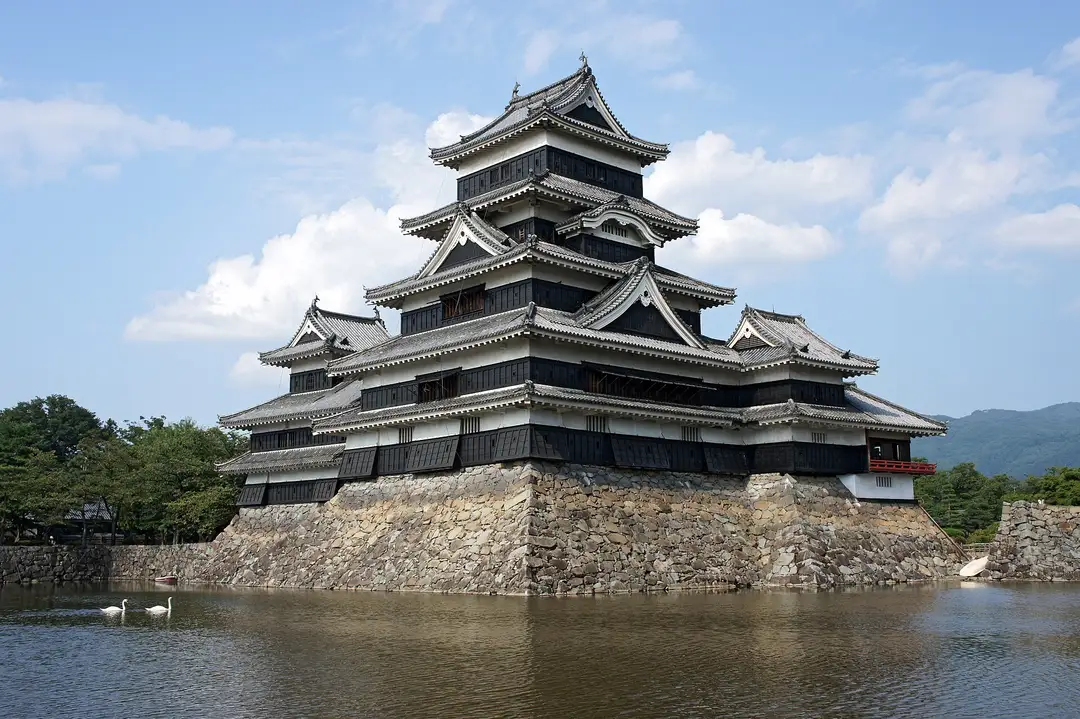 The black keep of Matsumoto Castle above its stone base and moat, with swans on the water and the Northern Alps behind
