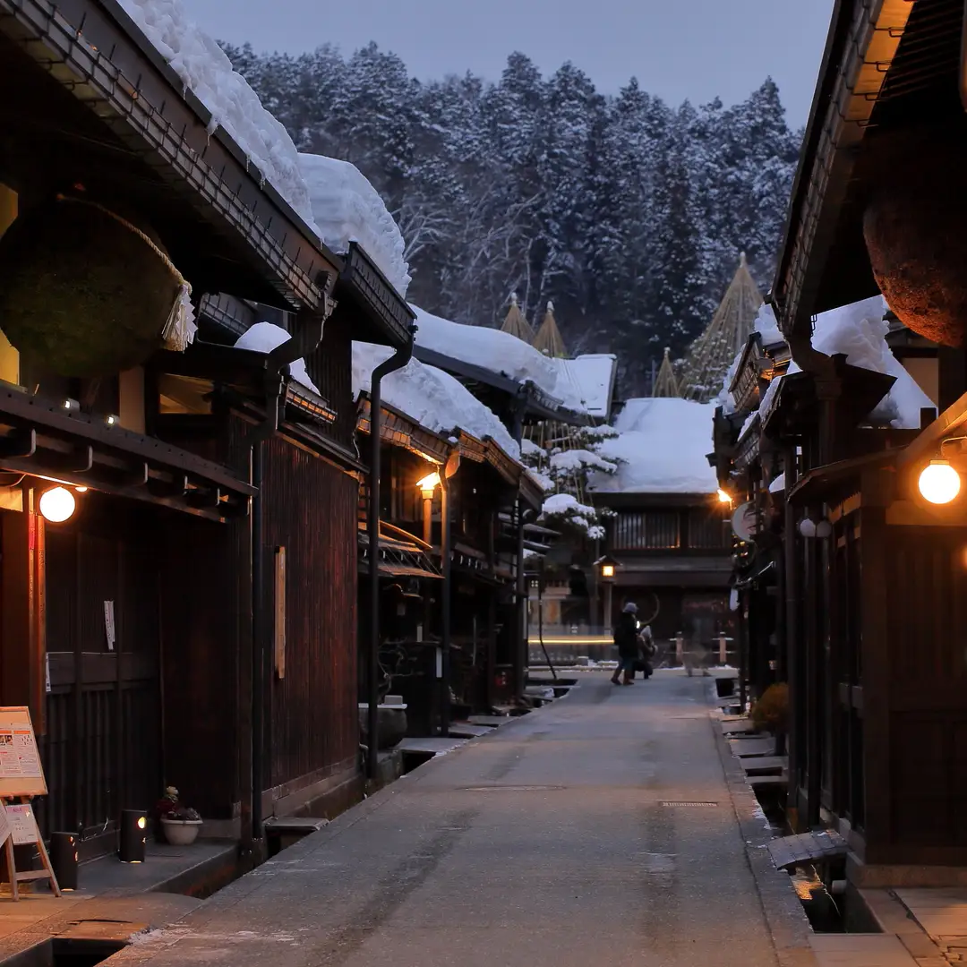 A dark-timber Sanmachi lane in Hida-Takayama at winter dusk, snow on the eaves and lanterns glowing