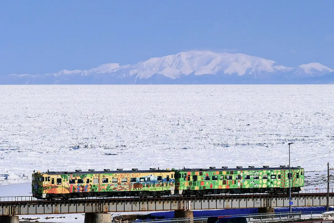 A local train crossing a bridge above a sea packed with drift ice, a snow-capped Hokkaido mountain beyond