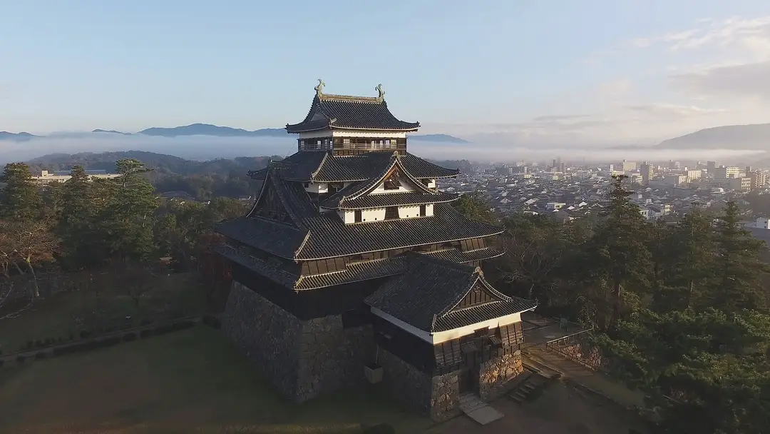 The black original keep of Matsue Castle rising above the misty town at dawn, golden shachihoko on its roof, forested hills and Sea-of-Japan haze beyond
