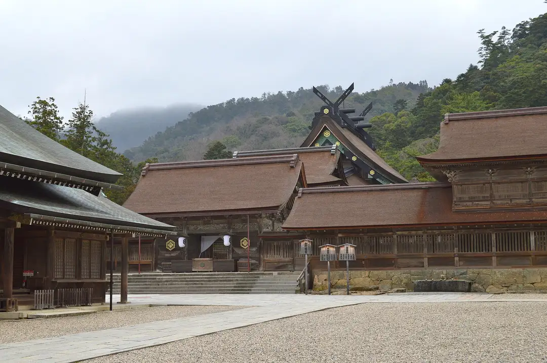 The cypress-bark roofs and crossed black chigi finials of the main sanctuary of Izumo Taisha, set against a misty forested hill above an empty gravel court