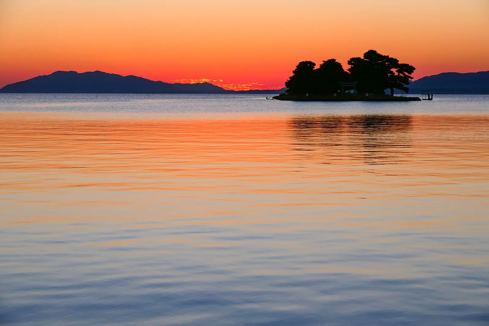 The wooded islet of Yomegashima silhouetted against a burning orange sunset over the mirror-calm water of Lake Shinji at Matsue, its small torii and pines dark against the sky