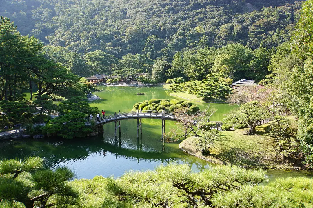 The arched Engetsukyō bridge reflected in the South Pond of Ritsurin Garden, hand-pruned pines and the wooded slope of Mt Shiun rising behind, Takamatsu