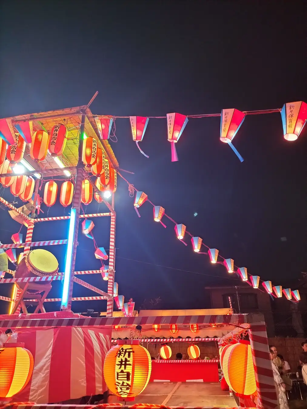 Strings of red-and-white paper lanterns and a drum-tower yagura lit up at night at a neighbourhood bon-odori