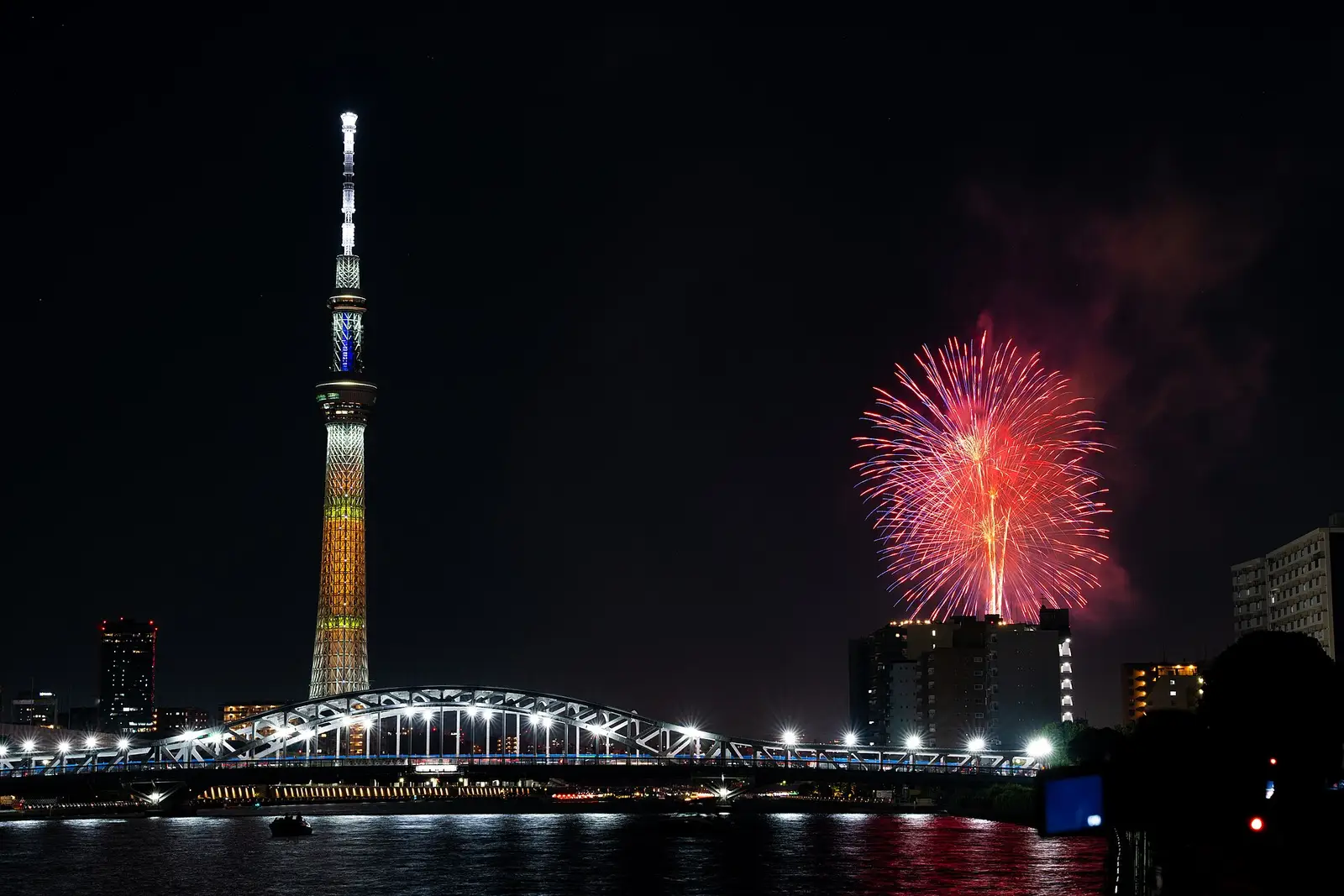 Fireworks blooming over a river at night during a Japanese summer hanabi festival, the illuminated Tokyo Skytree on the skyline