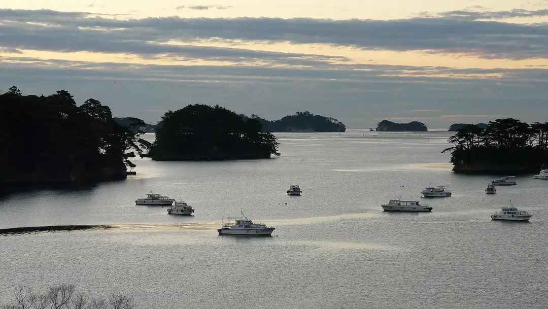 An elevated dusk view of Matsushima Bay, pine-clad islets scattered across calm mirror-like water with a few small tour boats