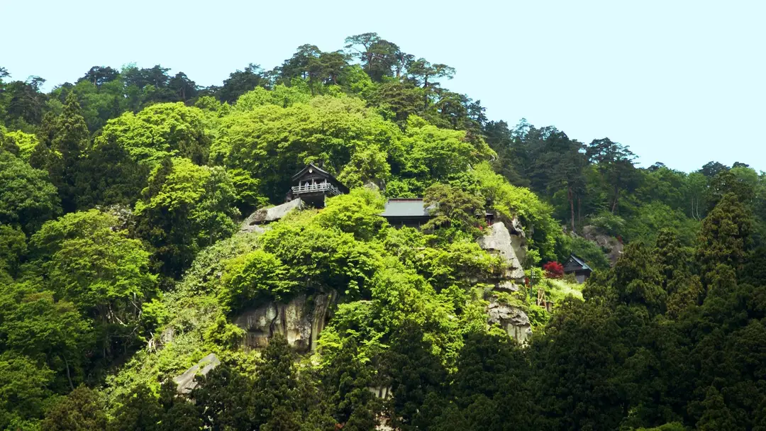 The wooden halls of Yamadera (Risshaku-ji) perched on a rocky cliff ledge amid dense green forest above the valley