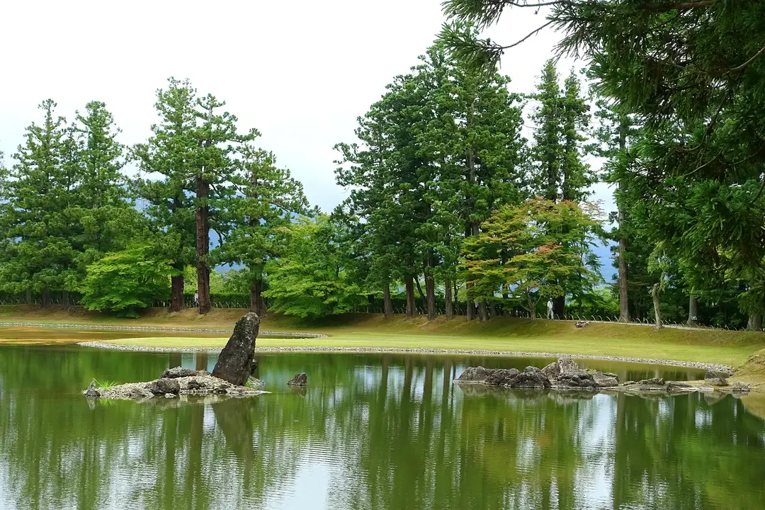 The still Ōizumi-ga-ike pond of Mōtsū-ji's Pure Land garden at Hiraizumi, an upright shoreline stone among rocks, ringed by trees reflected on the calm water