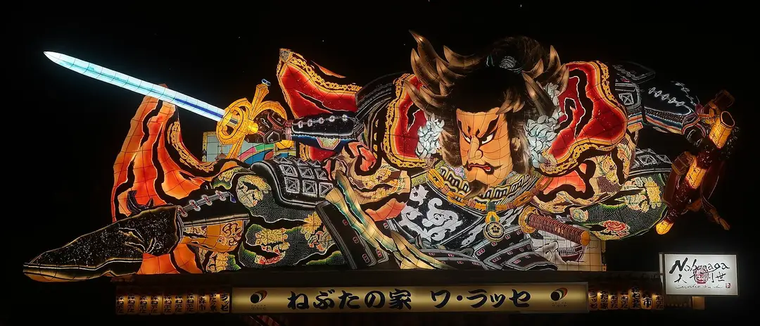 A giant illuminated Aomori Nebuta float of a fierce armored warrior brandishing a glowing blue sword, lit from within against the black night, filling the frame