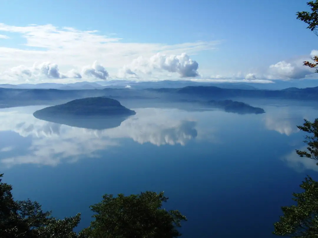 A wide elevated view of the deep-blue Lake Towada caldera from Mount Ohanabe, a forested peninsula and ringing hills mirrored in the calm water
